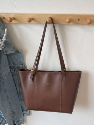 Brown structured tote bag made of PU leather with gold buckle detail and embossed logo.