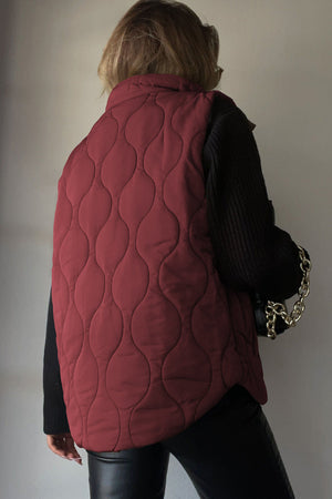 Wine quilted stand-collar gilet with wave pattern, worn over black sweater and pants.