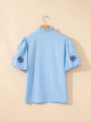 Light blue textured blouse with embroidered floral detail on puff sleeves and ruffled neckline.