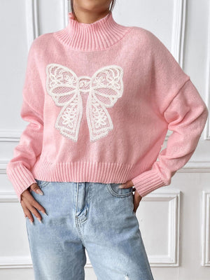 Pink knit turtleneck sweater with white lace bow appliqué detail.