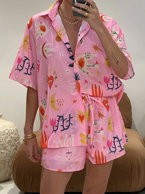 Pink co-ord set with short sleeves, button front, and drawstring shorts. Features a celestial and floral print.