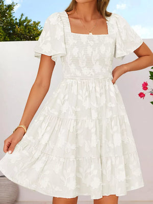 White smocked mini dress with tiered skirt, flutter sleeves, and floral embroidery.