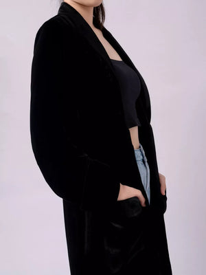 Black velvet longline trench coat with draped lapels and pockets.