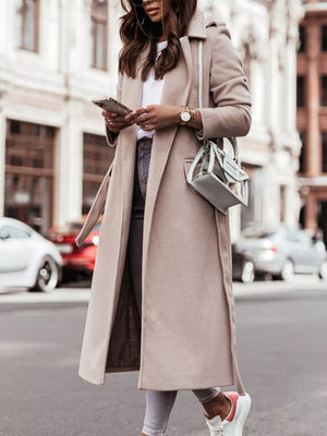 Belted longline trench coat in apricot with flap pockets and a tie belt, worn casually.