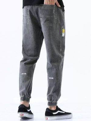 Men's grey relaxed fit drawstring jeans with elastic cuffs and subtle leg embroidery.