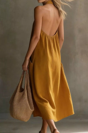 Mustard halter midi dress with pockets and open back, held by a model with a straw bag.
