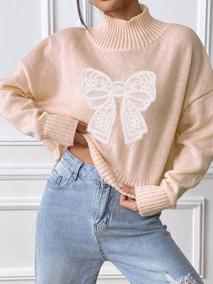 Blush pink turtleneck knit sweater with white lace bow appliqué detail.