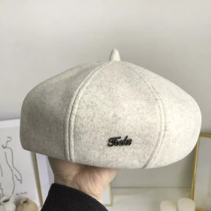 Ivory woolen beret with distinct paneled design and metal logo detail.