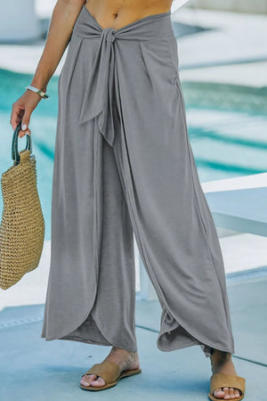 Gray wrap-front palazzo pants with slit hem and tie waist, styled with a straw bag.