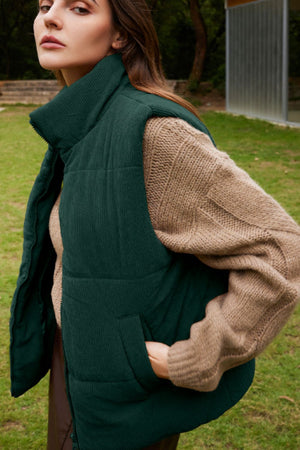 Dark green corduroy padded vest with a high collar and pockets, styled over a tan sweater.
