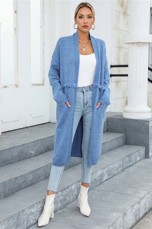 Light blue longline knit cardigan with fringe trim and pockets, worn open over a white top and jeans.