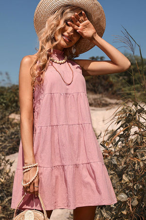 Pink halter neckline tiered mini dress with a bow-back detail, styled with a straw hat and jewelry.