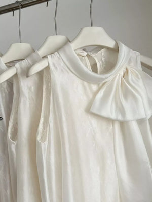 Sleeveless white shirt with a large bow detail at the neckline and subtle floral pattern.