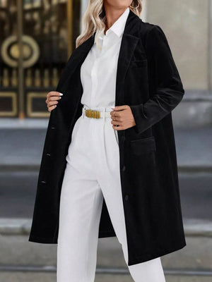 Black longline trench coat with lapel collar and buttoned front, worn open over white shirt and pants.