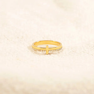 Gold pavé cross band ring with clear stones on a textured white surface.