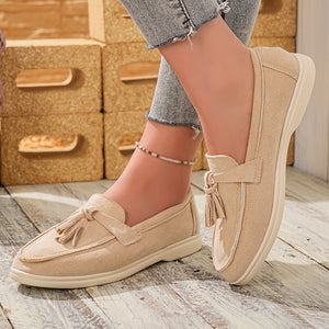 Beige faux-suede slip-on loafers with decorative tassels and a comfortable flat sole.
