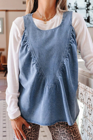 V-shaped pleated denim sleeveless top with a rounded neckline, styled over a white long-sleeve shirt.