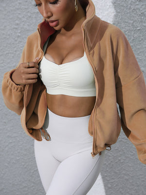 Tan fleece cropped funnel neck jacket with zipper detail, worn open over a white ruched sports bra.