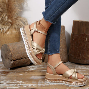 Gold bow knot wedge sandals with ankle buckle and braided trim detail.