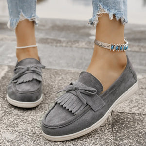 Gray suede loafer flats with fringe bow detail and white soles.