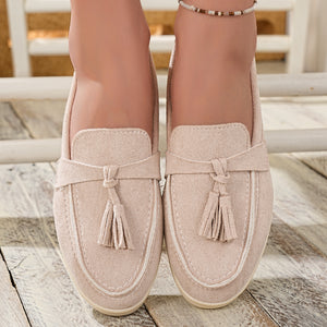 Beige faux-suede slip-on loafers with decorative tassels and contrast stitching.