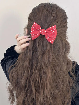 Deep red polka dot bow hair clip with white dots, styled in wavy brown hair.