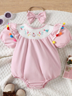 Blush pink cotton baby romper with smocked birthday motif neckline and pom-pom trim.