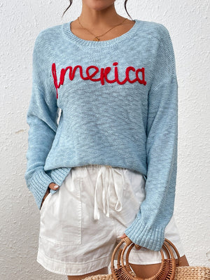 Pastel blue knit sweater with red embroidered "America" text, crew neck, long sleeves.