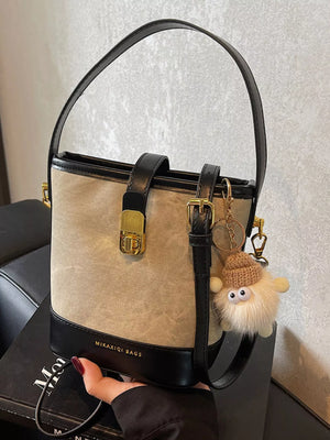 Elegant bucket bag in tan canvas with black PU leather trim and gold hardware, featuring a fuzzy charm.