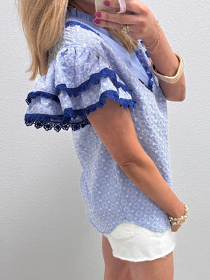 Light blue eyelet embroidered blouse with flutter sleeves and navy lace trim.