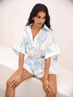 White linen romper with blue cocktail print, tie waist, and buttoned shirt top.
