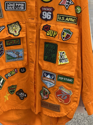 Orange denim trucker jacket with assorted embroidered patches and buttoned chest pockets.