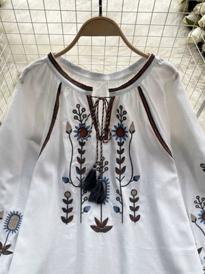 White blouse with tie neck, billowy sleeves, and colorful floral embroidery.