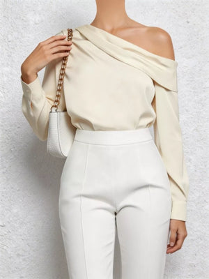 Cream one-shoulder draped blouse with a long sleeve, styled with white pants and a chain strap purse.