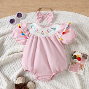 Blush pink baby romper with smocked birthday motif neckline, puff sleeves, and colorful pom-pom trim.