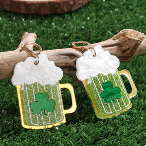 Gold dangle earrings shaped like beer mugs with white foam, green glitter, and clover charms.