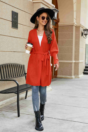 Orange-red knit cardigan with puffed sleeves and a tie waist, worn open over a white top.