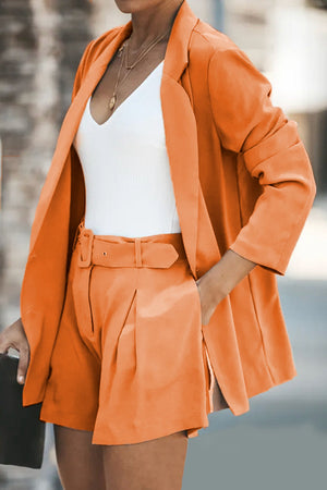 Orange double-breasted blazer and pleated shorts set with a white V-neck top and gold necklaces.