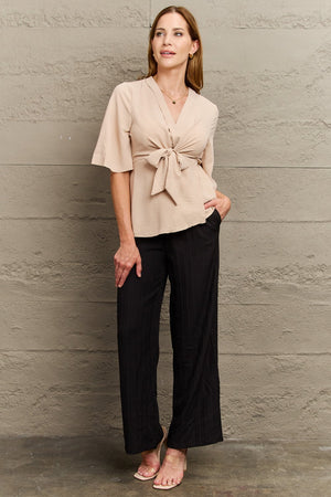 Light apricot wrap-front blouse with tie waist and flutter sleeves, styled with black pants.