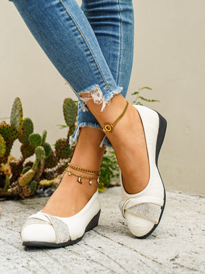 White slip-on wedge shoes with rhinestone bow detail and low wedge heel.