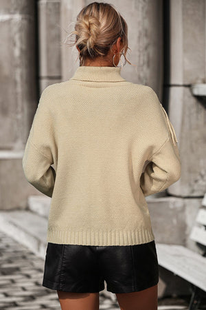 Beige knit turtleneck sweater with fringe detail on sleeves, textured knit.