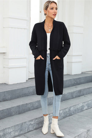 Black longline knit cardigan with fringe trim and pockets, worn open over a white top.