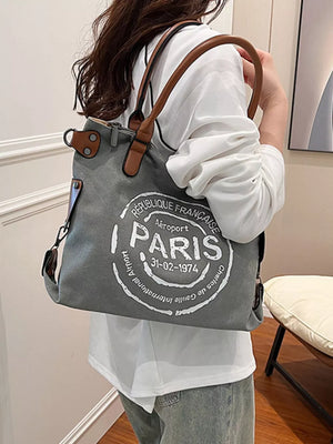Parisian graphic tote bag in dark gray canvas with brown handles and airport stamp detail.
