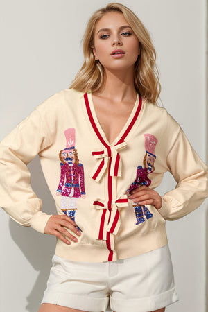 Cream knit cardigan with sequin nutcracker embellishments, red trim, and bow tie closures.