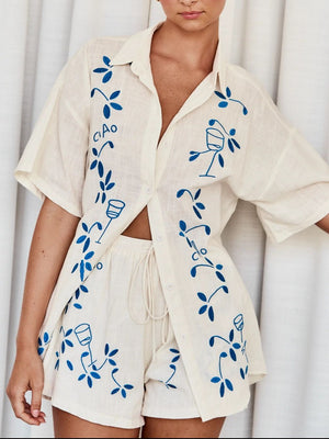 Cream linen co-ord set with blue embroidery of leaves and wine glasses, featuring a button-up shirt and drawstring shorts.