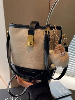 Elegant bucket bag in tan canvas with black PU leather trim and gold hardware, with a fluffy charm.