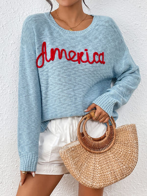 Pastel blue knit sweater with red embroidered "America" text, crew neck, long sleeves.