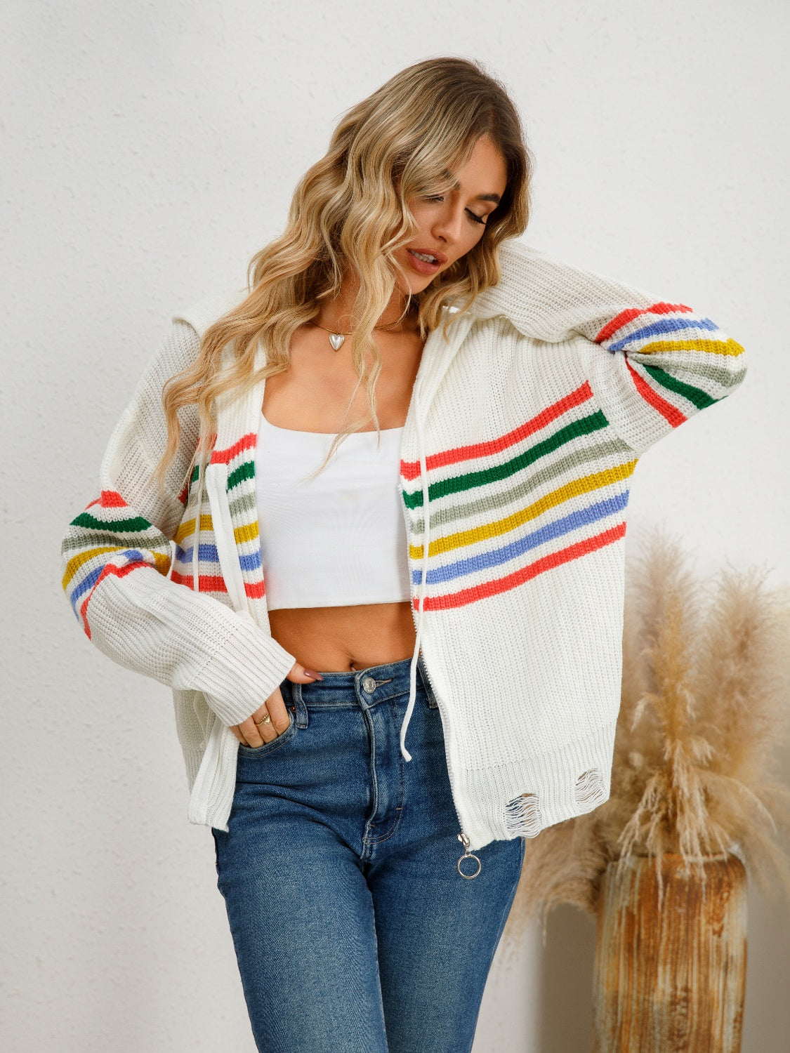 White distressed knit cardigan with rainbow stripes on sleeves and front, featuring a zipper closure.