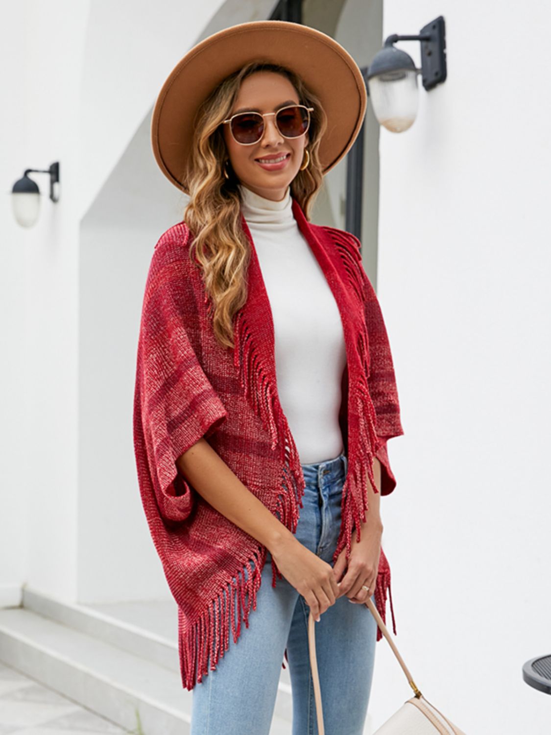 Textured red fringe capelet with striped pattern and open front, styled with a white turtleneck and jeans.
