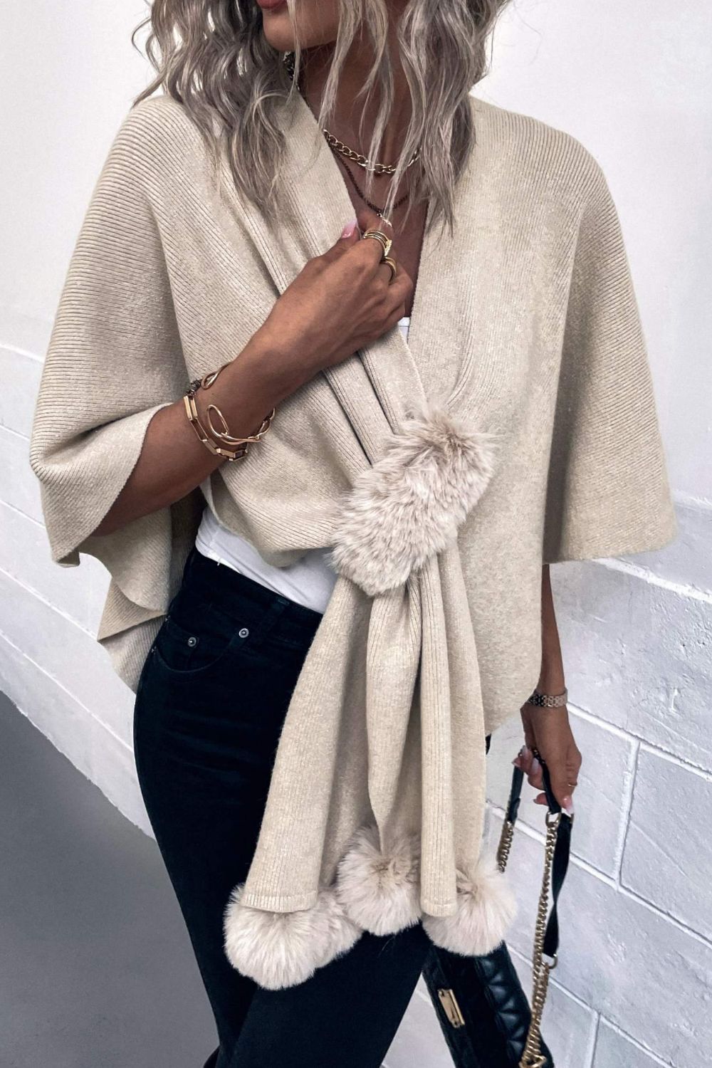 Taupe knit cape with fur trim and faux fur pom-poms, draped open over a white top.